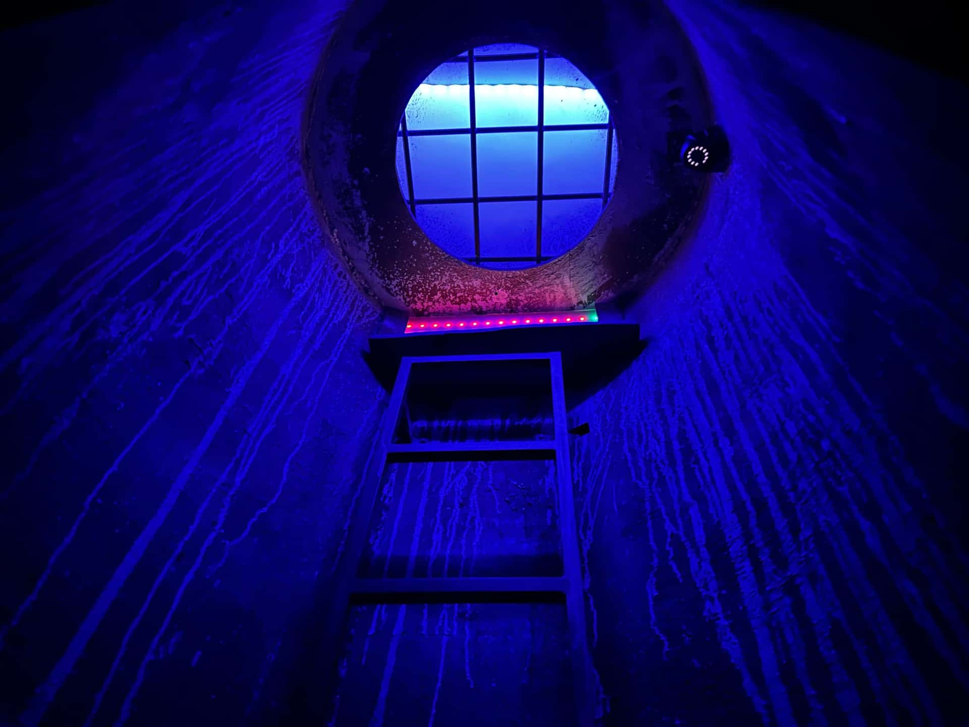 Blau beleuchteter Schacht mit Leiter und rundem Gitterfenster im Escape Room Robot Paranoia in Rosenheim